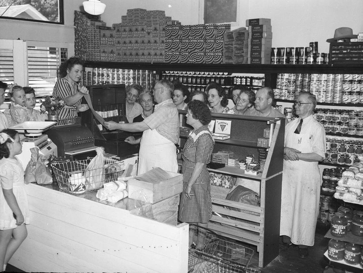 May 28, 1945: Trimble Grocery and Market at 1615 Cooper St. has been a fixture in Fort Worth for 17 years. Owner E. Burt Trimble is selling to his market associate, Walter Jetton (second from right). Mr. Trimble plans enjoy retirement and take a much needed rest. Customers, neighbors, and friends are gathering in the store to pay their respects to the retiring grocer. His wife is behind him. Standing on a box by the “McCaskey” bookkeeping system is Mrs. C.B. Rains. She is presenting a scroll, which all of Mr. Trimble’s customers expect to sign by next week. (The store building no longer exists at Cooper and 8th Avenue, near the Cook Children’s campus.)