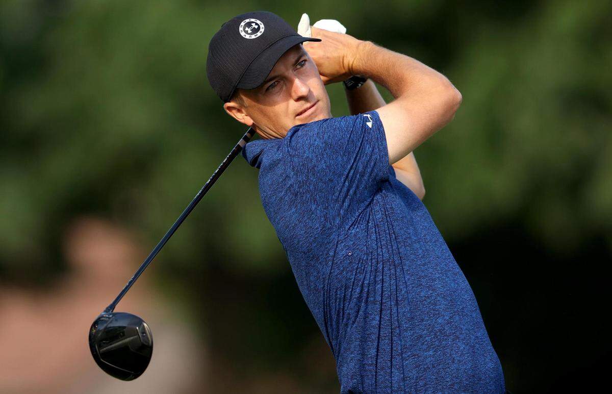 A man wearing a dark blue shirt and black baseball hat looks off to the right while at the end of his golf swing.