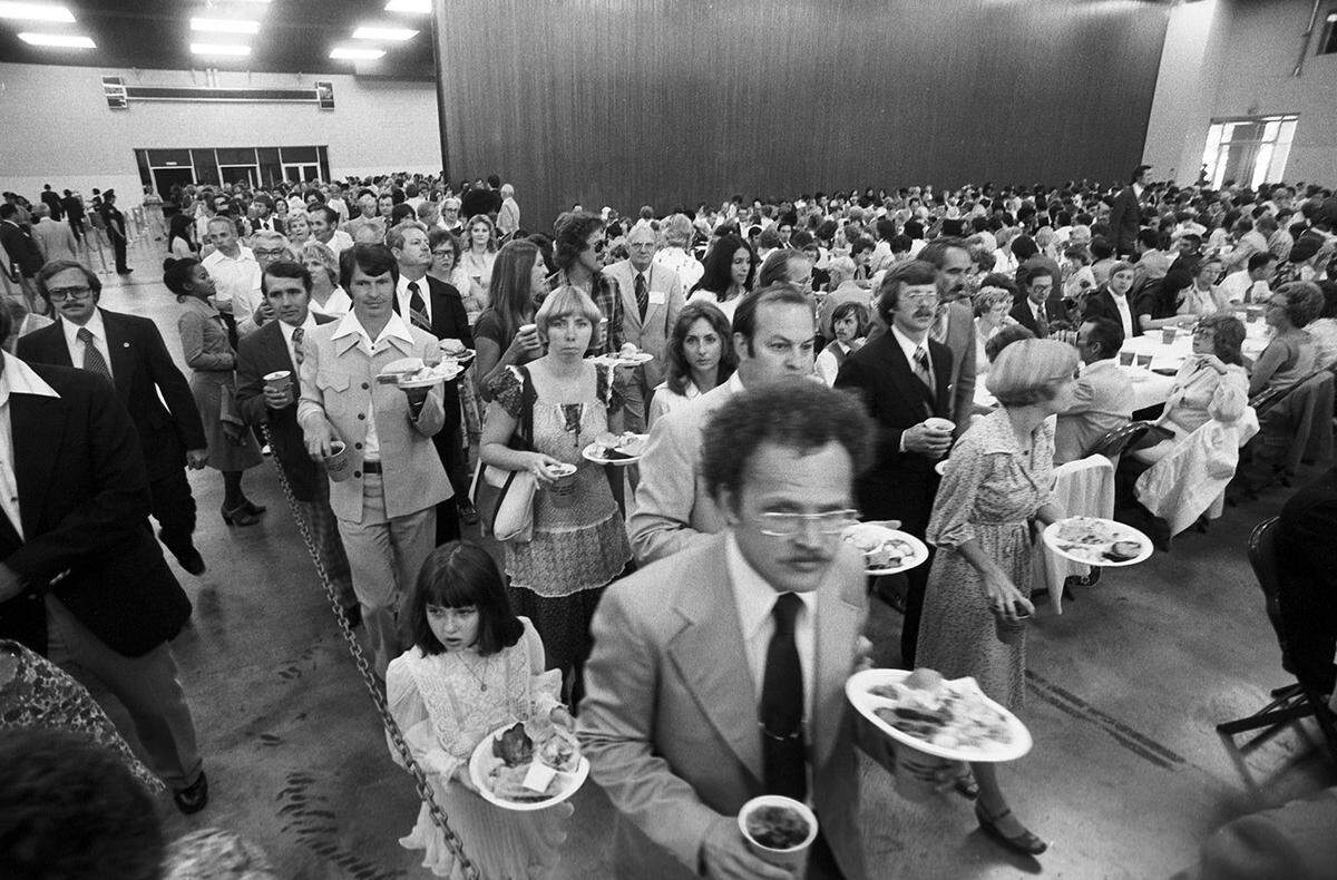 A line of people including a few children, holding plates of food, with a large crowd of seated people at the right.