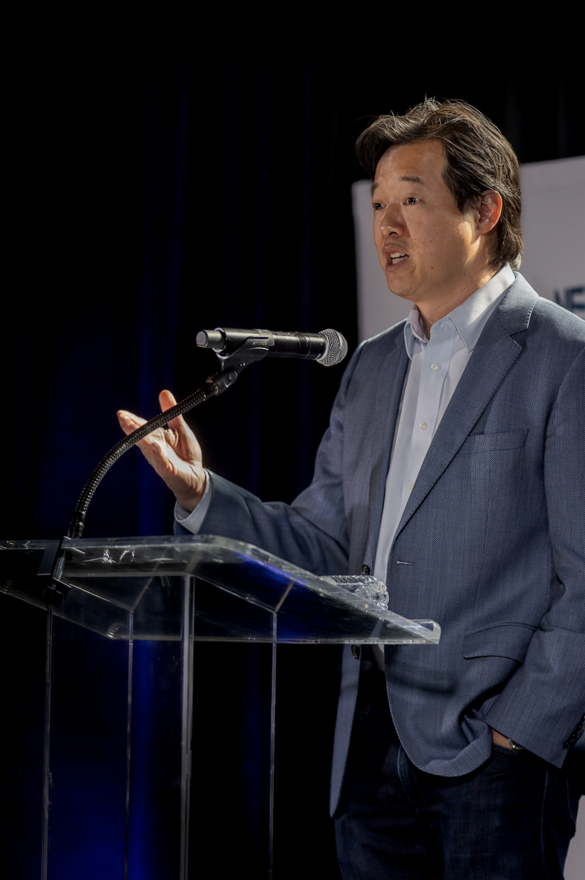 Victor Hwang wears a blue business suit and stands at a glass podium with a microphone. His left hand is in his pocket while his right hand is held up in a gesturing position as he delivers his speech.