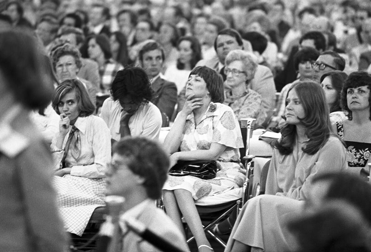 A seated crowd listens to the speech, with one woman holding her chin as she appears to listen intently.