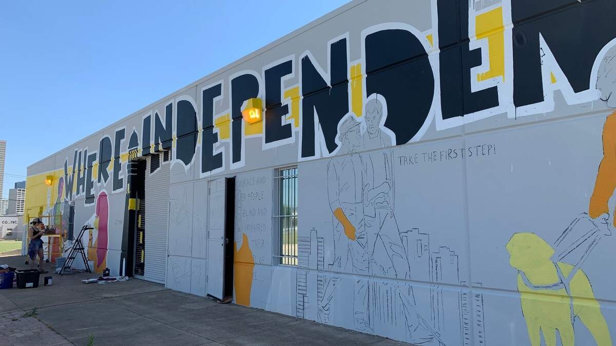 Artist Kristen Soble is pictured to the left, standing in front of a painted gray wall with black words toward the top of the wall that read "Where independence begins". A lighthouse has been painted in between the words "where" and "independence" and there are other partially finished images in various colors depicting blind people living every day life, such as walking with a guide dog, reading and cooking.