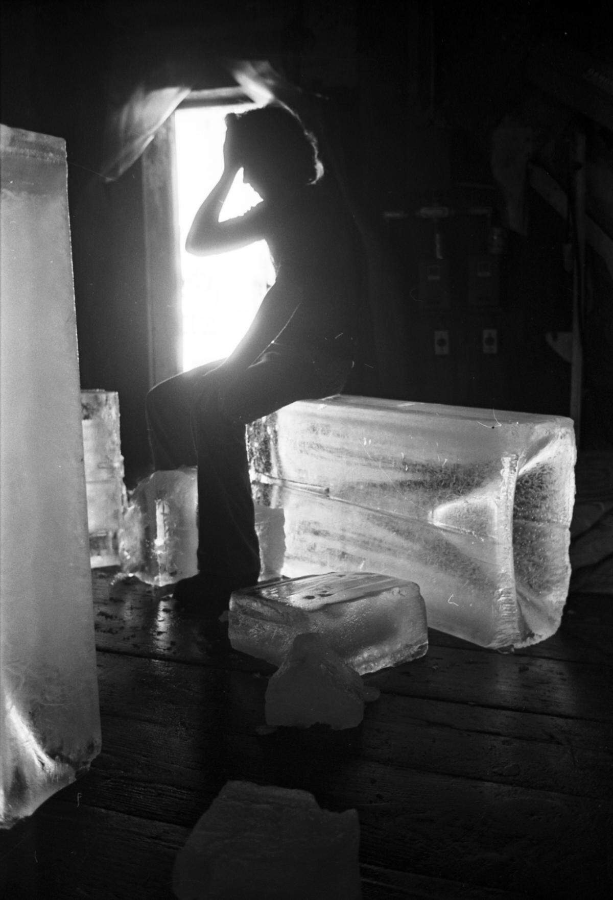 The man, in silhouette near a window, is sitting on a large block of ice.