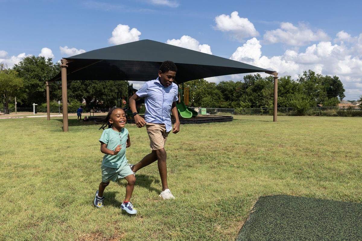 Two young boys, the boy on the left much shorter, run across a grass space with a playground canopy in the background.