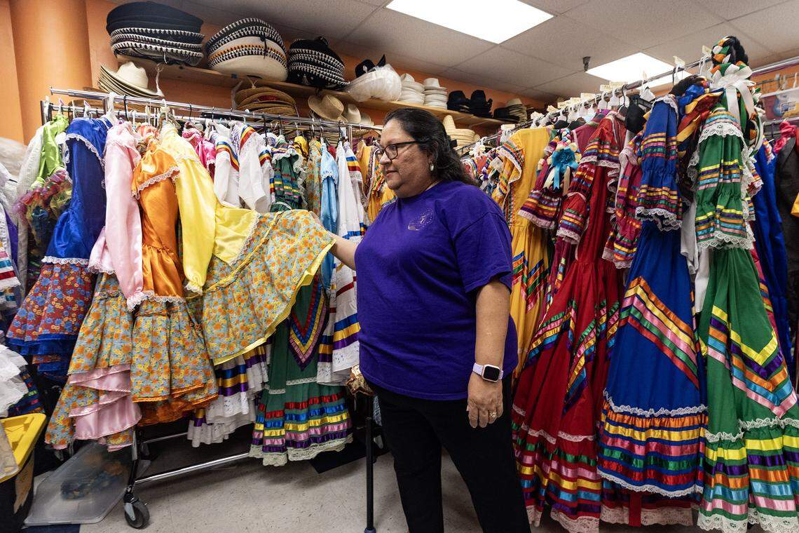 Carol Alvarado lifts the skirt of many vibrantly colored dresses and costumes hanging on racks.