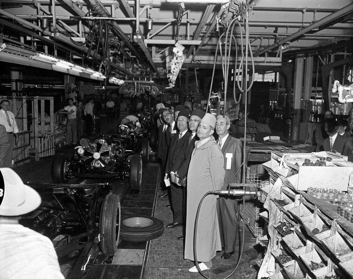 The king and several others, all wearing robes and fezzes, look at vehicle chassis rolling down an assembly line. 