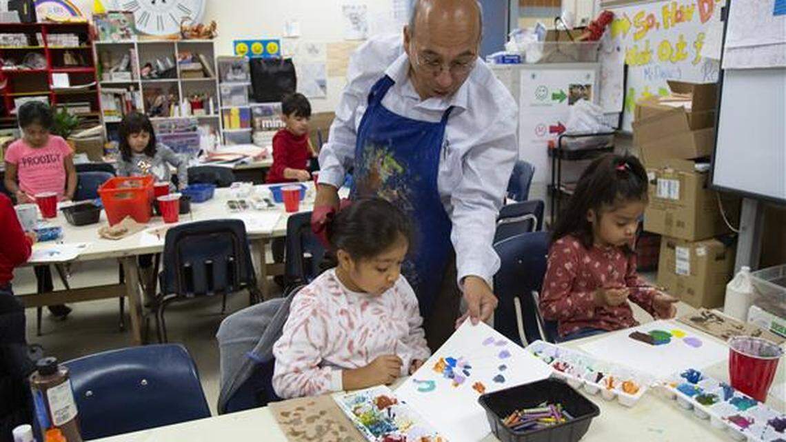 A man in a blue apron leans down and picks up a painting done by a girl sitting at a table. Nearby, another girl works on a painting.