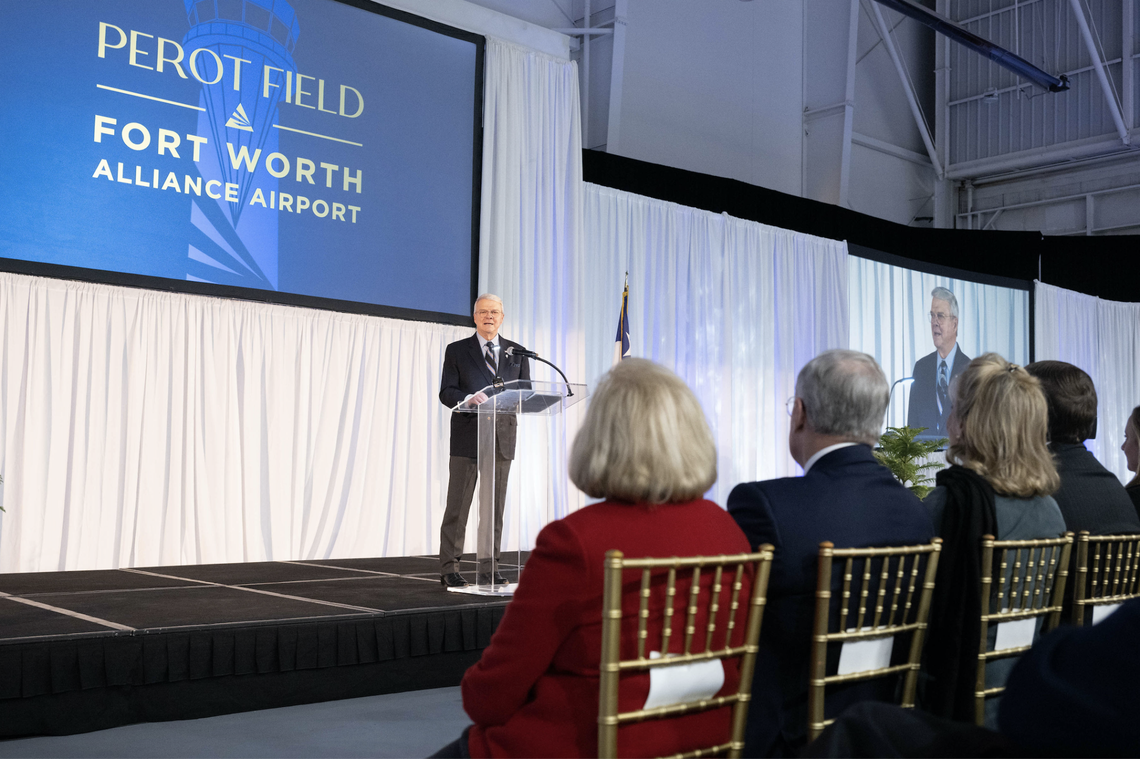 A zoomed out photo of McArtor shows him standing at a glass podium in a business suit in front of a row of event guests sitting in chairs. Guests are dressed in business attire. McArtor stands in front of a presentation screen reading "Perot Field Fort Worth Alliance Airport," large white curtains and an American flag.