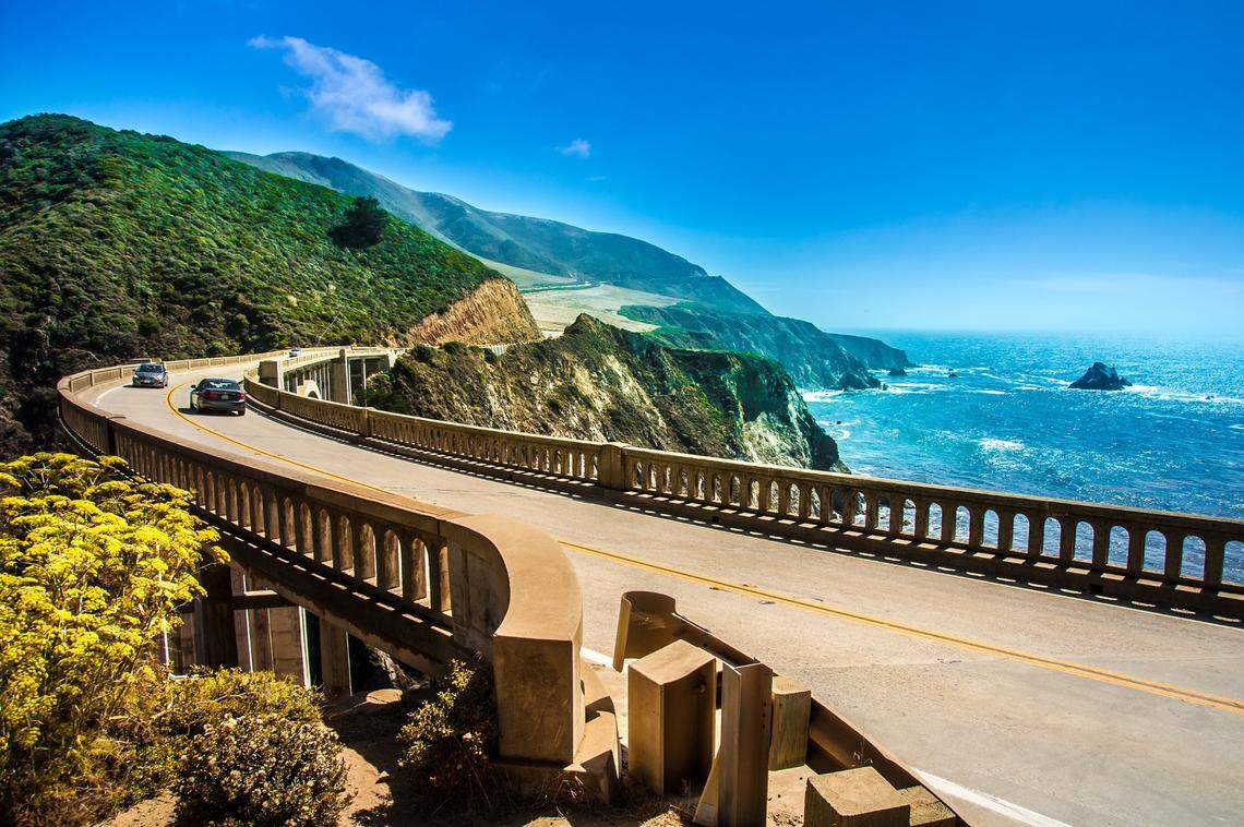 A winding highway rests along the coast of California. To the right of the two-lane road is the vibrant, blue ocean and sky. To the left of the road is green hillside.