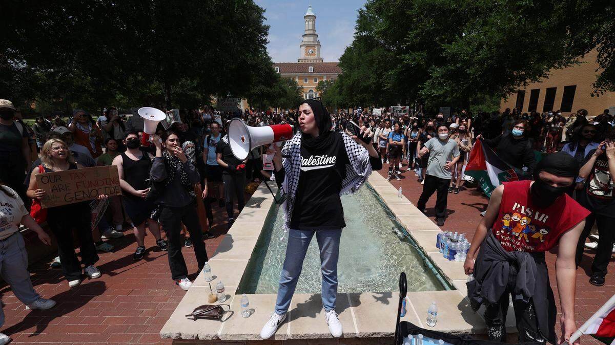 A person stands in front of a long rectangular fountain, shouting into a bullhorn, while surrounded by people, some with signs. 