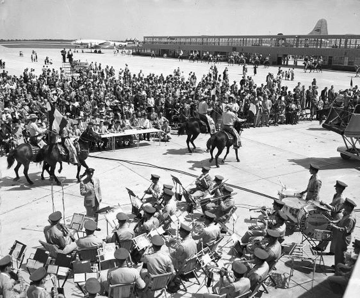 A large number of spectators are standing or seated in chairs watching the proceedings. In the foreground is the Carswell Air Force Base Band. In the distance there are two parked airplanes. The airport is also known as the Greater Southwest International Airport and Amon Carter Field.
