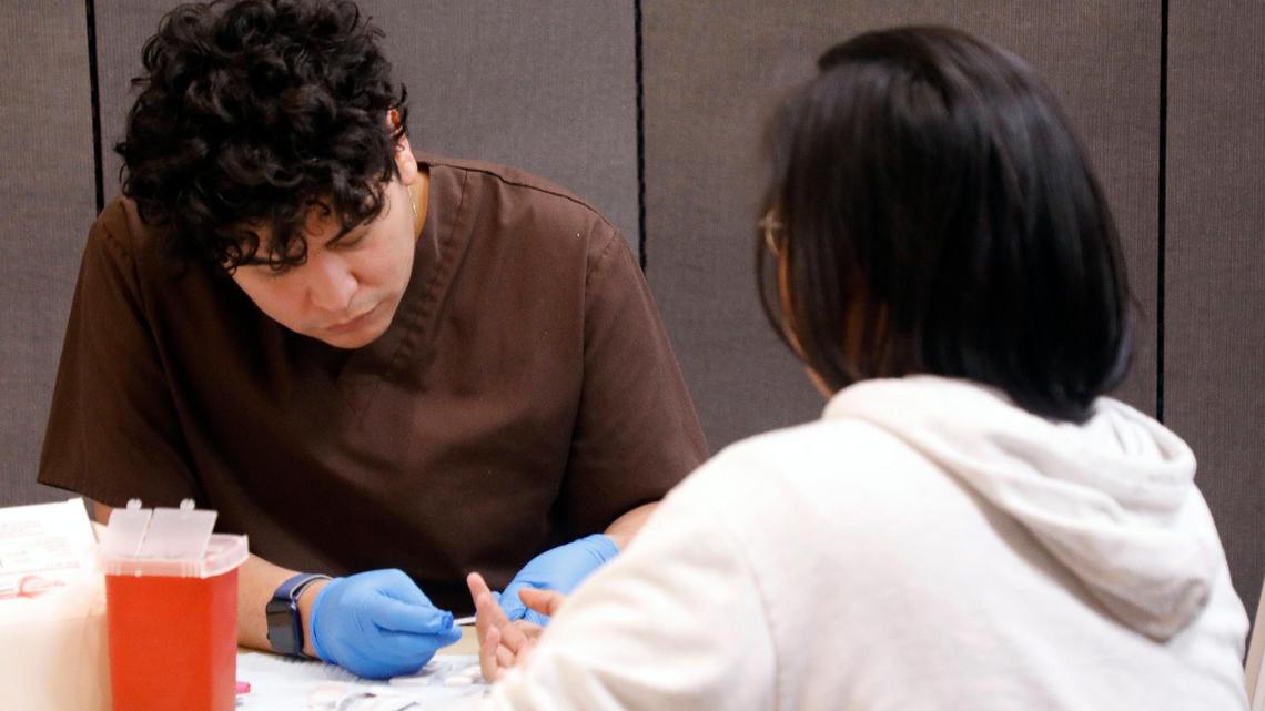 A health worker in a brown sweatshirt leans over a woman's hand as he prepares to prick her fingerpad for blood. 