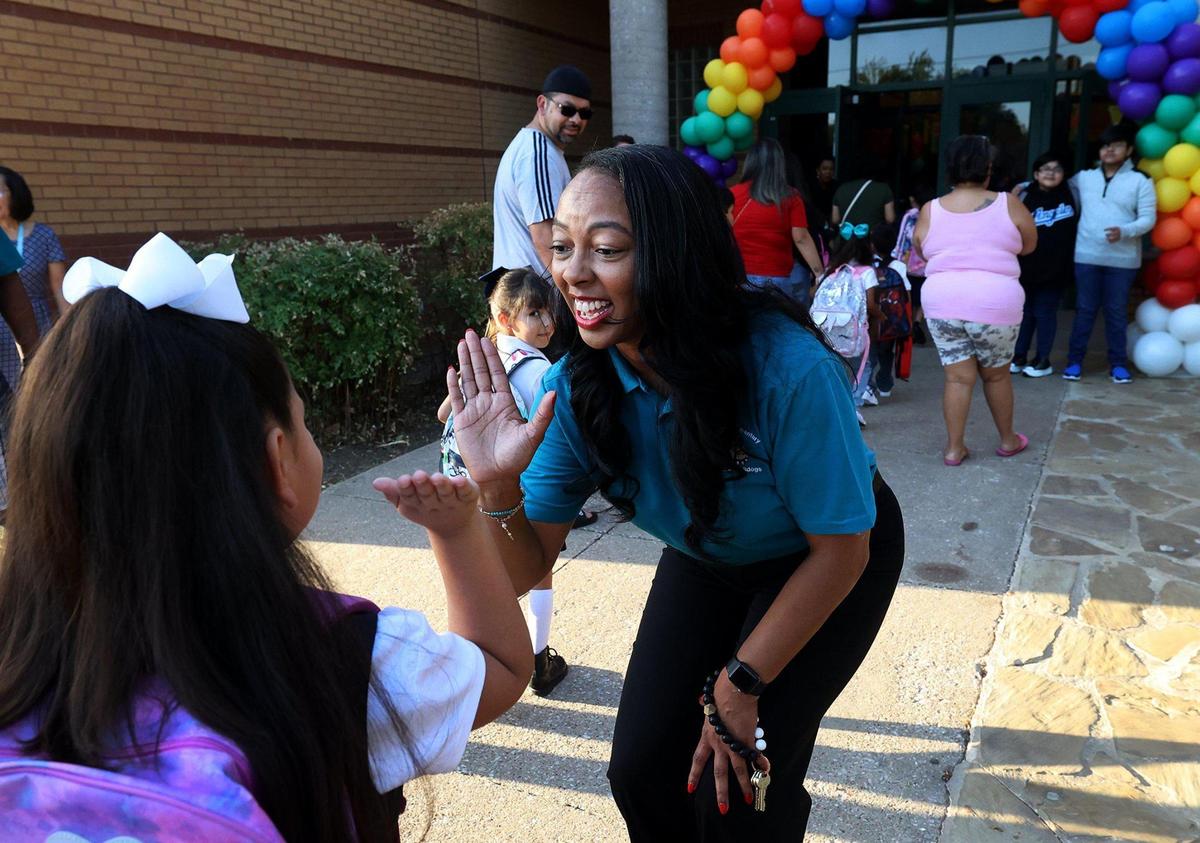 Woman high fives a small child with a bow in her hair in front of the school, which is decorated with balloons. 