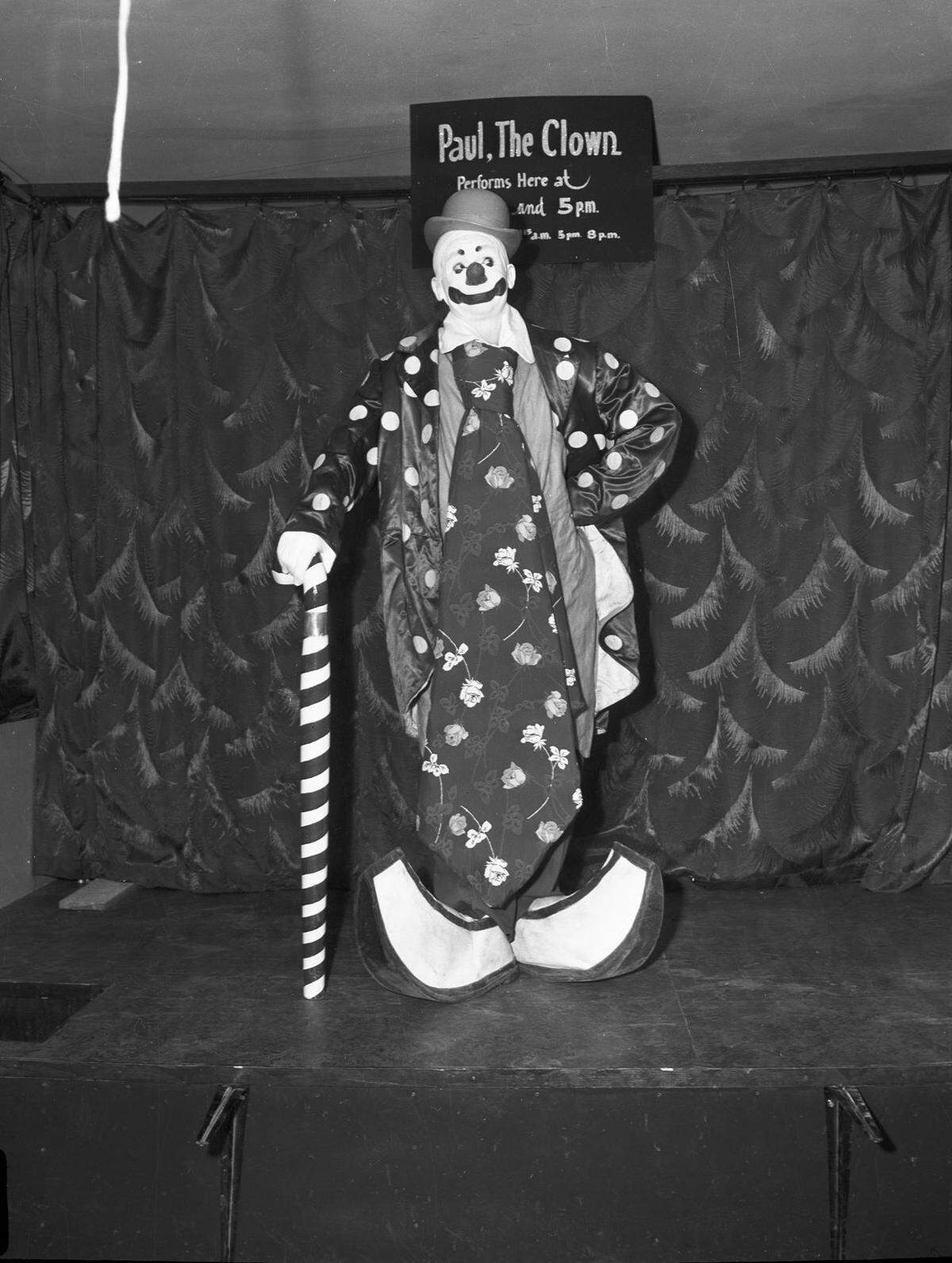 A clown poses with huge oversized shoes holding a giant candy cane.