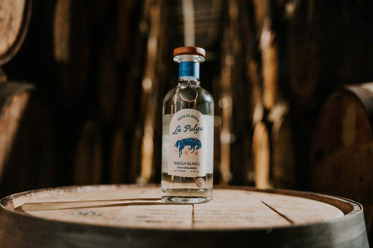 A glass bottle propped on a wooden barrel with the La Pulga label facing forward. The label reads, "Hecho en Mexico, La Pulga, Tequila Blanco" and has an image of a bucking bronco.