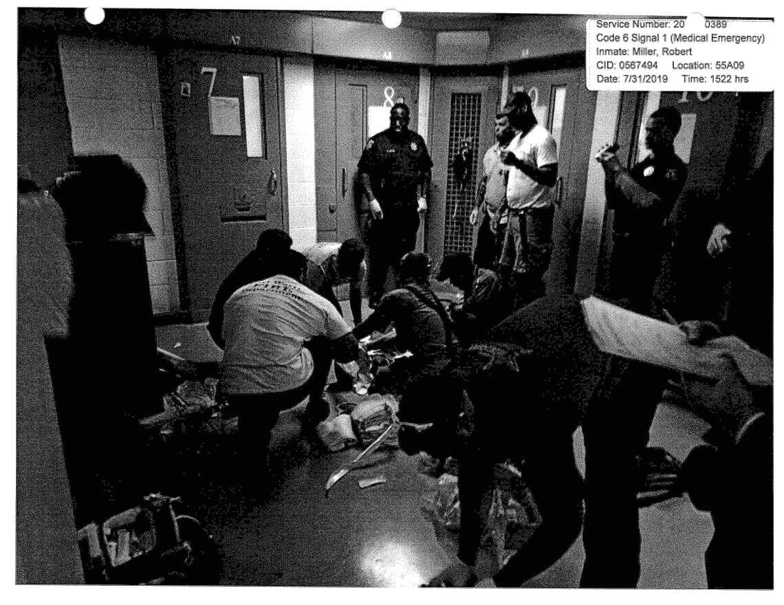 A black and white photo shows several medics huddled around Miller on the ground, while jail officers and others stand around and watch. Miller isn't visible.