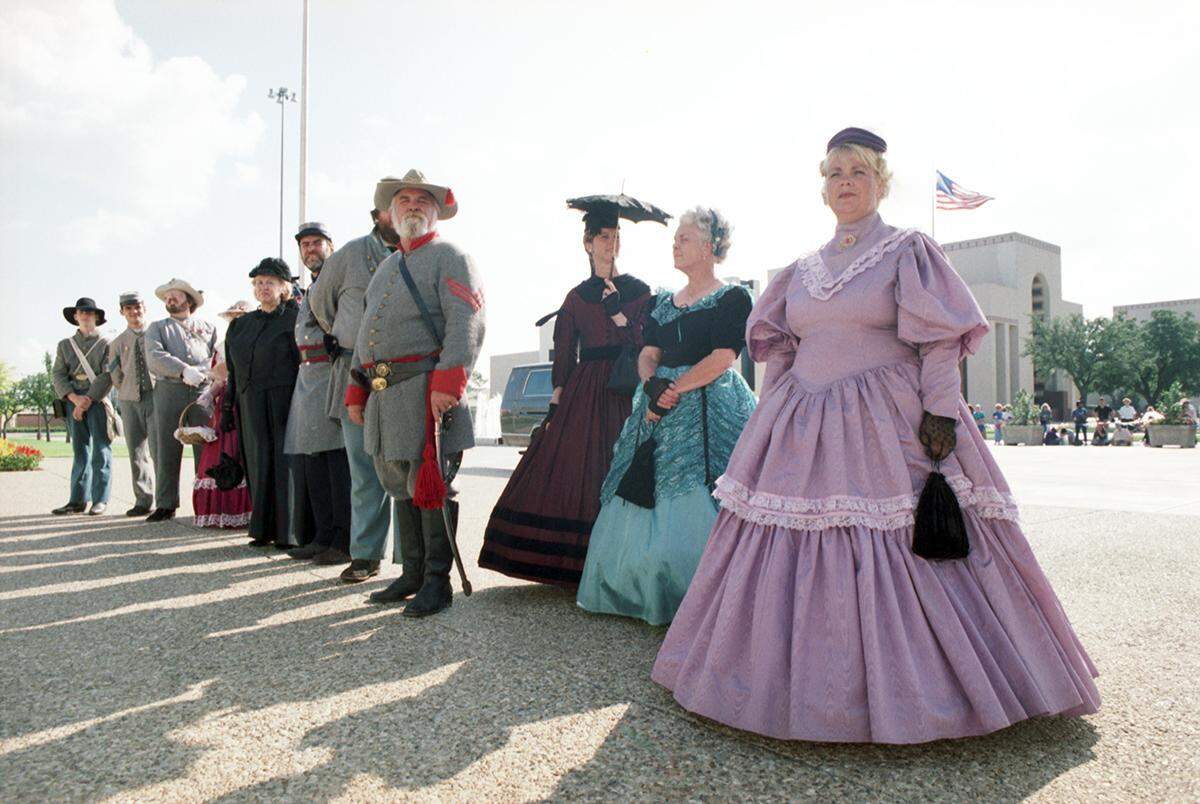 A line of people including women in a large colorful hoop skirts from the 1800s, and men dressed in Civil War uniforms, stand outside the Hall of State in downtown Dallas in honor of Queen Elizabeth's visit in 1991.