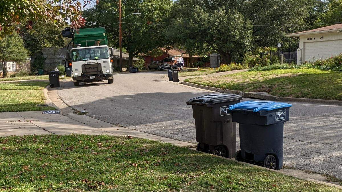 A garbage truck in the distance picking up trash in a tree lined street with two cans in the foreground.