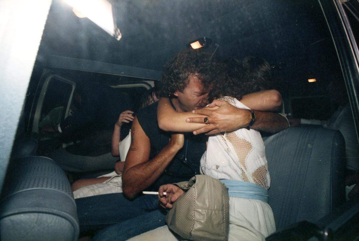 A photograph taken through a vehicle window that shows an emotional man and woman hugging.