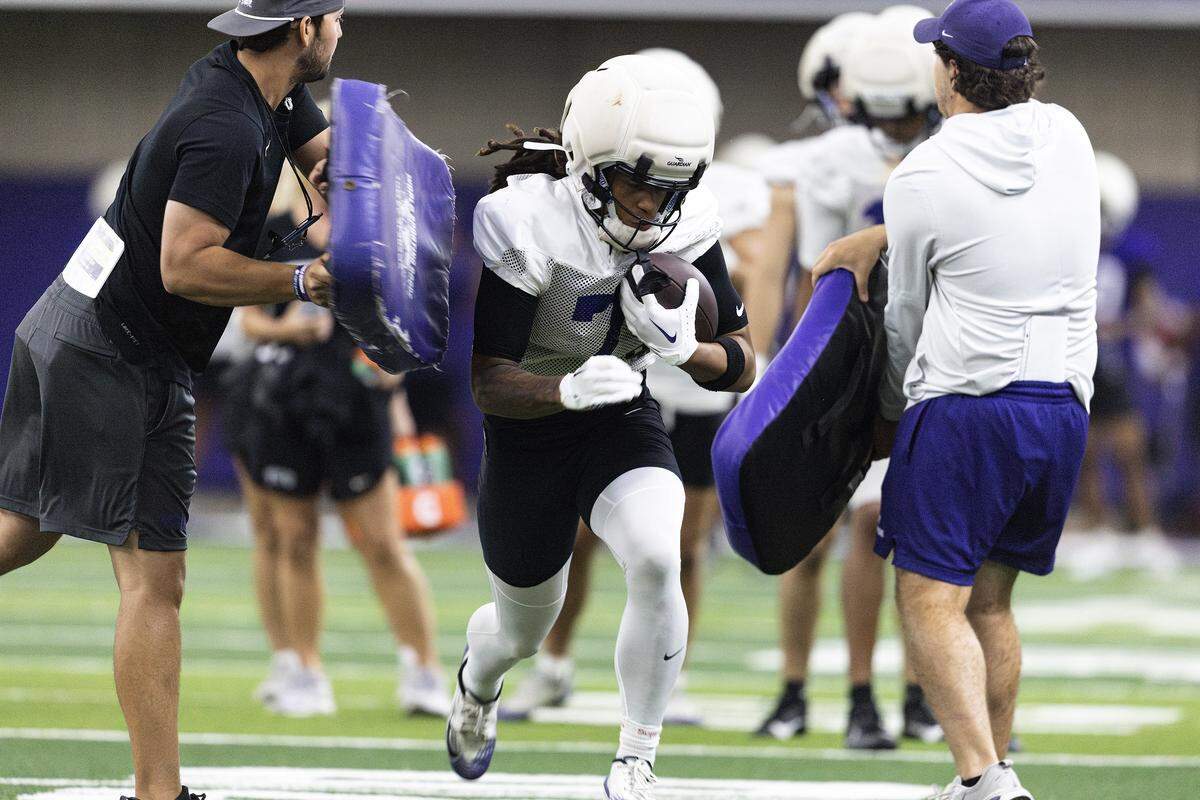 A football player runs through coaches holding purple pads on either side of him.