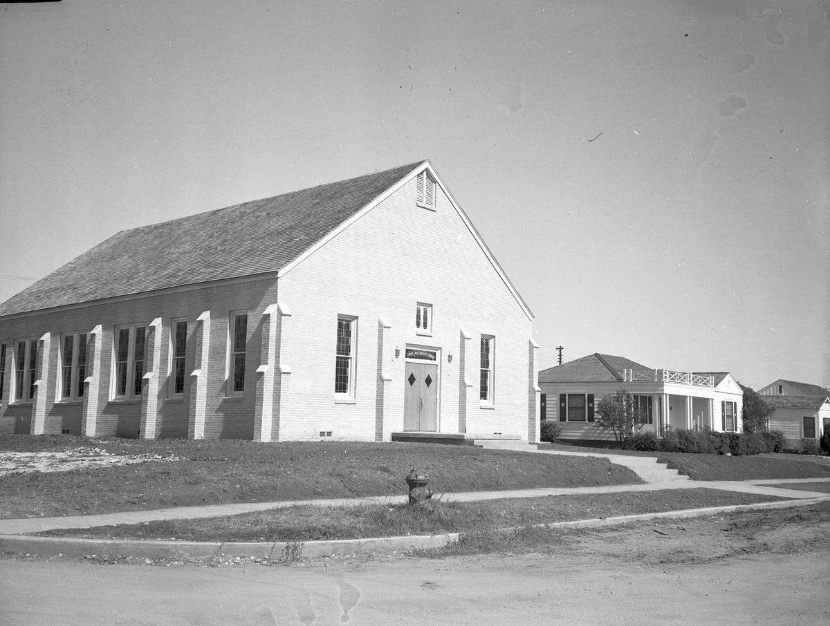 Grace Methodist Church. Signs of construction such as bare soil and gravel surround a modest brick church building at the corner of Calmont Avenue and Clover Lane in West Fort Worth. There are architectural features in the brick work that reference the buttresses found on large churches and cathedrals, there are ornamental leaded glass windows and the name of the church is placed in a glass piece over the double doors into the building. Next door to the church is a modest light colored house with an ornamental rail above the small front porch. Shutters embellish the windows and a rough hedge stands in front of the building. This is the parsonage. Clipping: "Its Dedication Is Today. Grace Methodist Church at Calmont and Clover Lane will be dedicated Sunday. At right is the parsonage, occupied by Rev. W. F. Smith.
