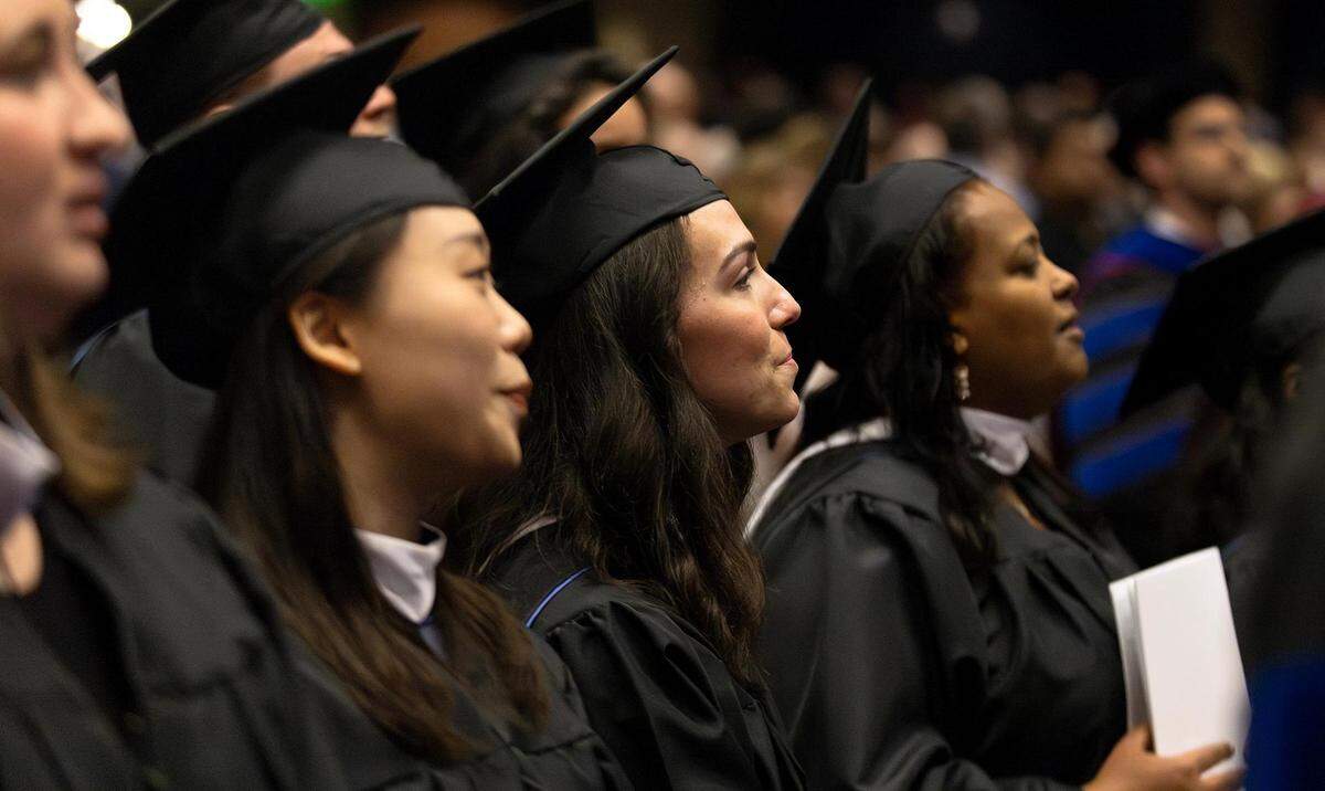 A line of graduates, wearing black cap and gown,  in profile.