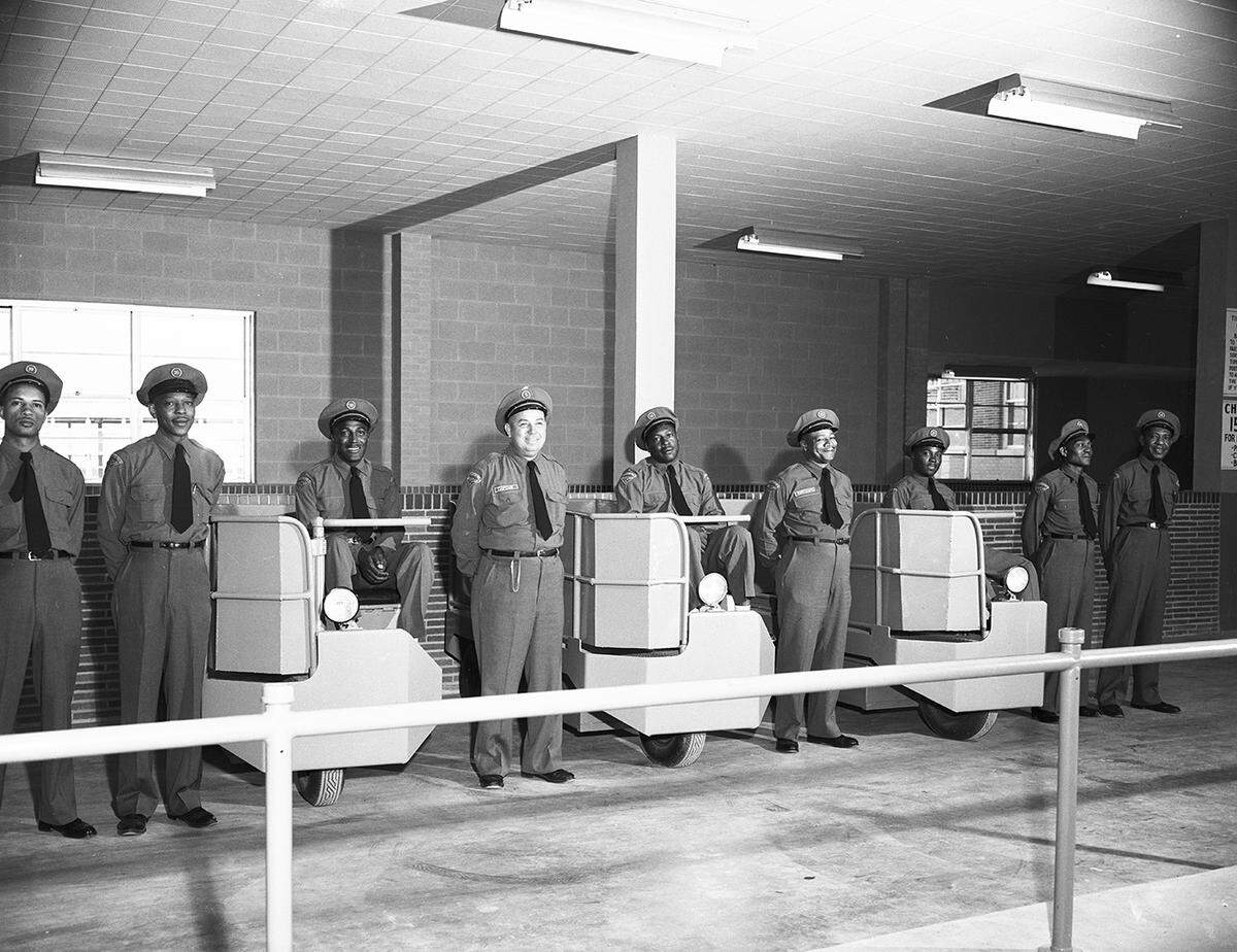 A row of smiling men in uniforms and hats pose for a camera. Three of the men are seated on wheeled carts. 