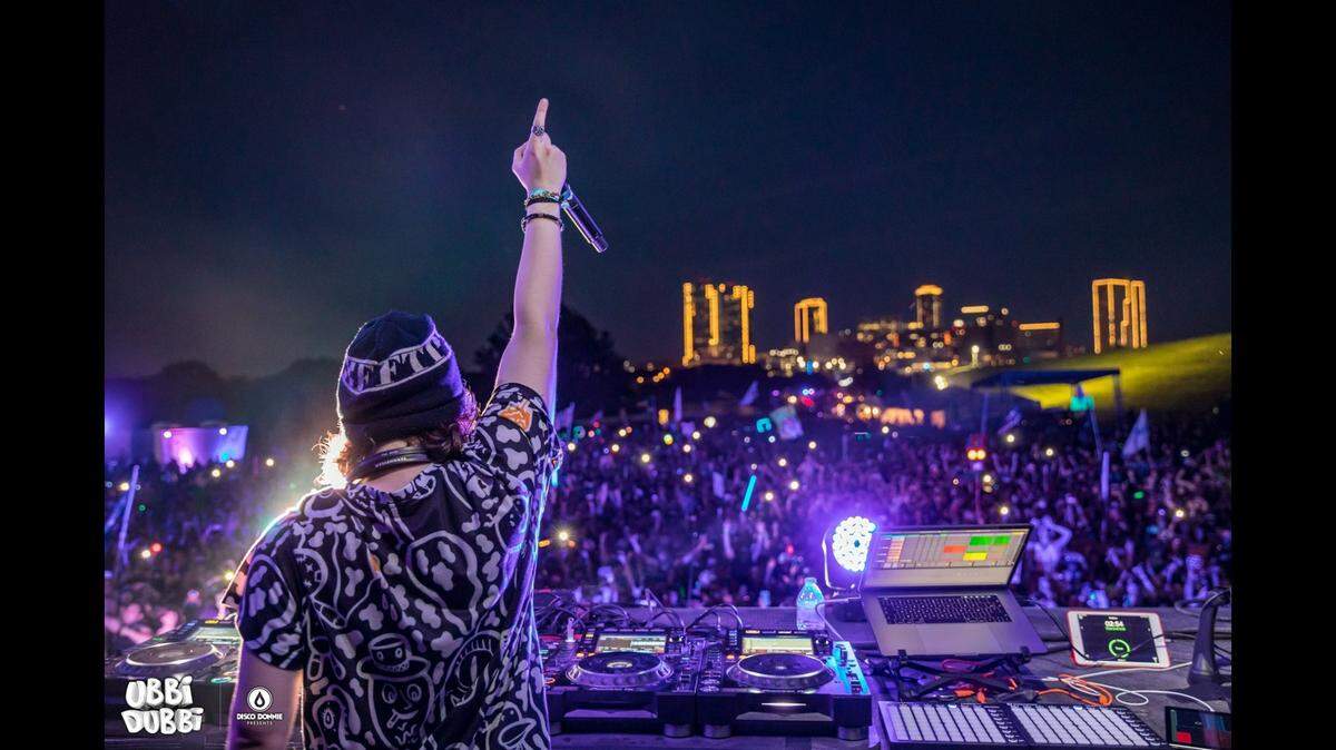A disc jockey from behind holding up a microphone to a large audience at Fort Worth's Panther Island Pavilion with the Fort Worth skyline in the background