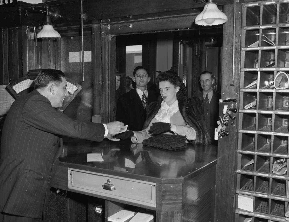 Miss Garland is wearing a bow in her hair, a suit with lace around the collar, and a fur jacket over her shoulders. Mr. Harris is dressed in a double breasted pinstripe suit. A crowd of people are standing in the background, watching the exchange. 