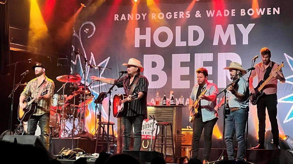 Two male lead singers and a backing band of three other men perform on stage with guitars in front of a backdrop that reads quote Randy Rogers and Wade Bowen: Hold My Beer.