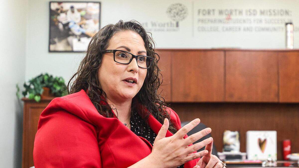 A woman in a red suit gestures with her hands while speaking. She sits in an office with the Fort Worth ISD logo on a wall behind her.