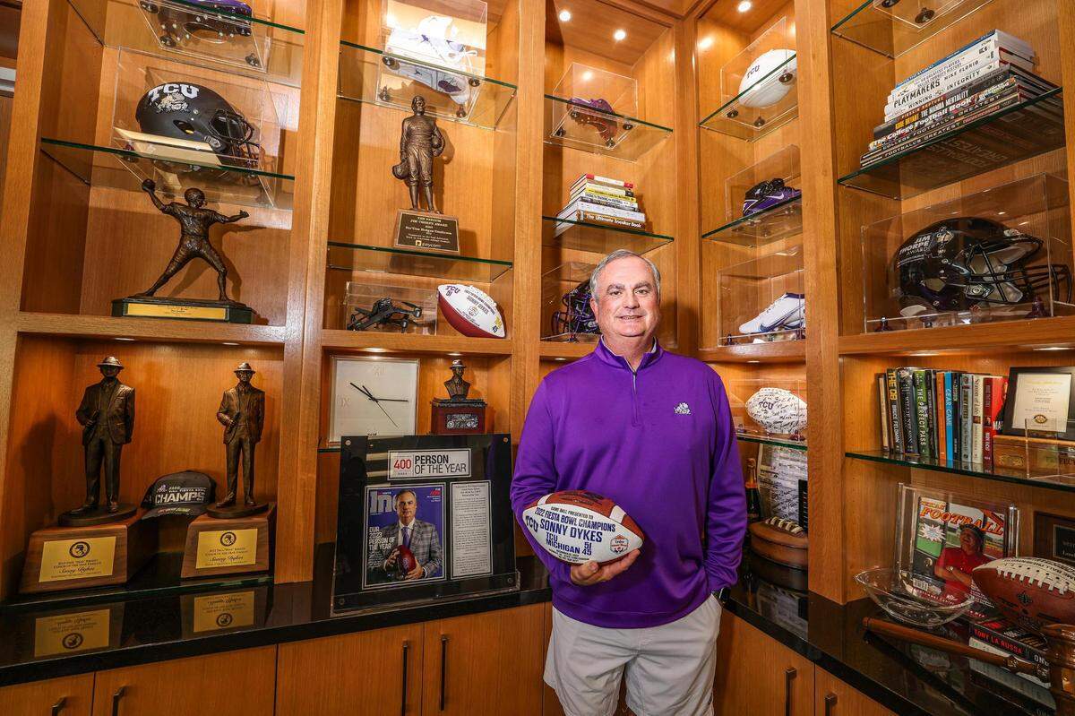 Sonny Dykes holds a National Championship football while standing in front of shelves filled with awards, trophies and book.