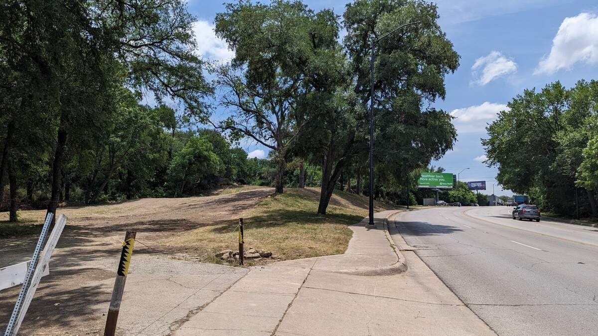 An empty plot of land next to a busy, tree-lined roadway. 
