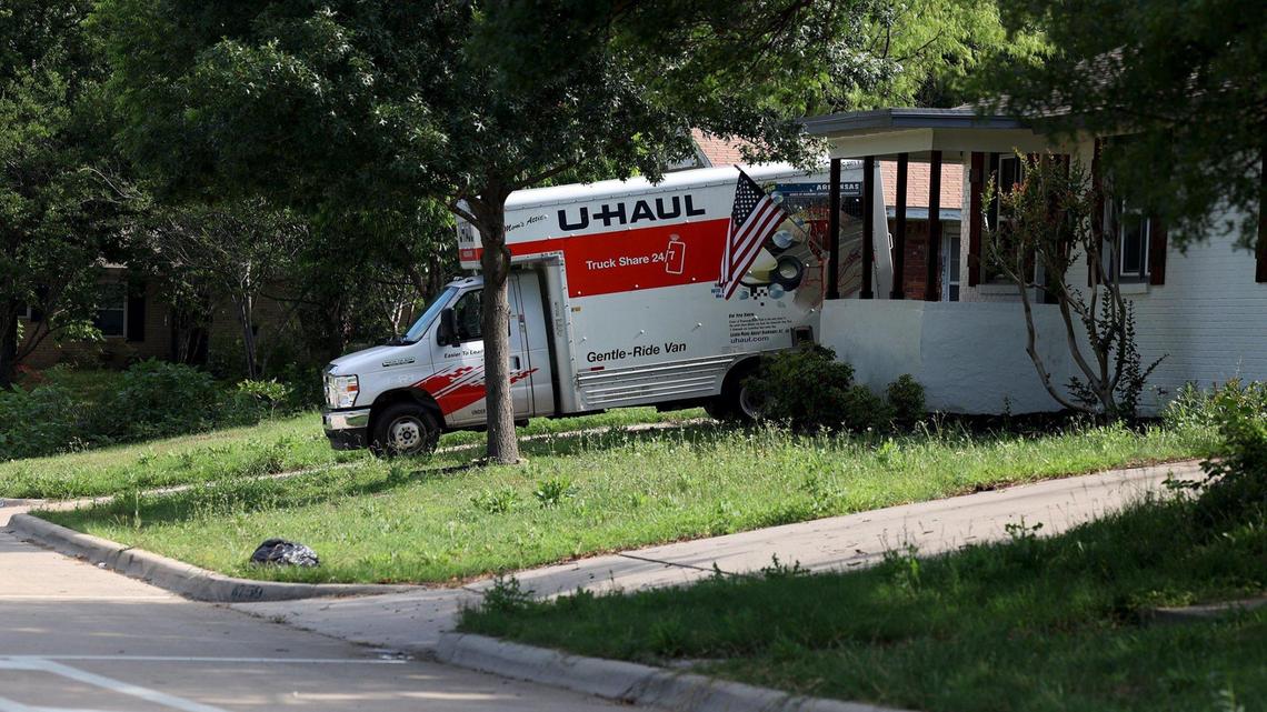 A red and white U-Haul truck backed into a white home's drive way, surrounded green grass and trees.