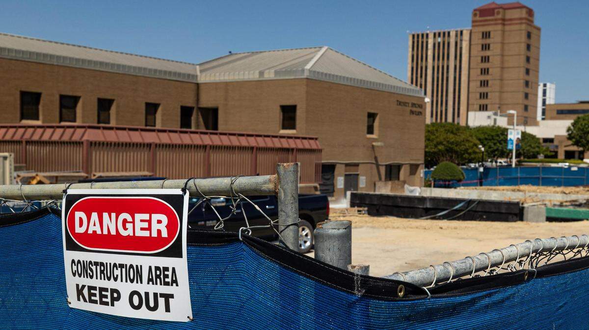 A sign that says ' Danger: Construction Area. Keep Out' is attached to a fence lined with a blue tarp. In the background you can see a dirt lot and buildints.