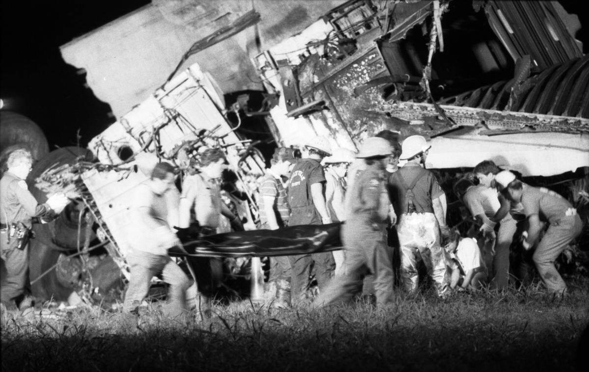Black and white photo of numerous emergency workers. In the background is  alarge section of plane wreckage.
