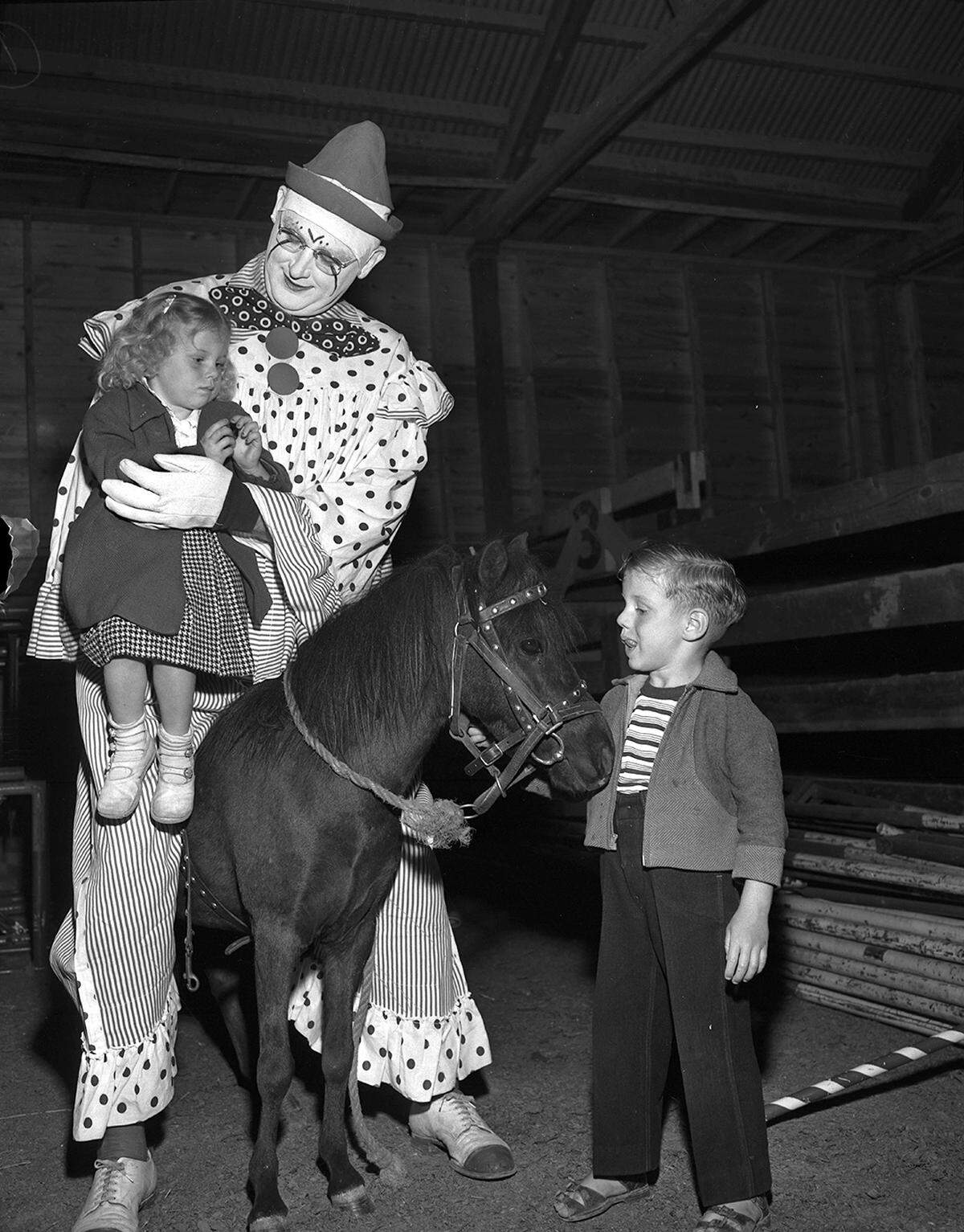A large man dressed like a clown on a horse holds a little girl who has a blank look on her face while a little boy stares at the horse.