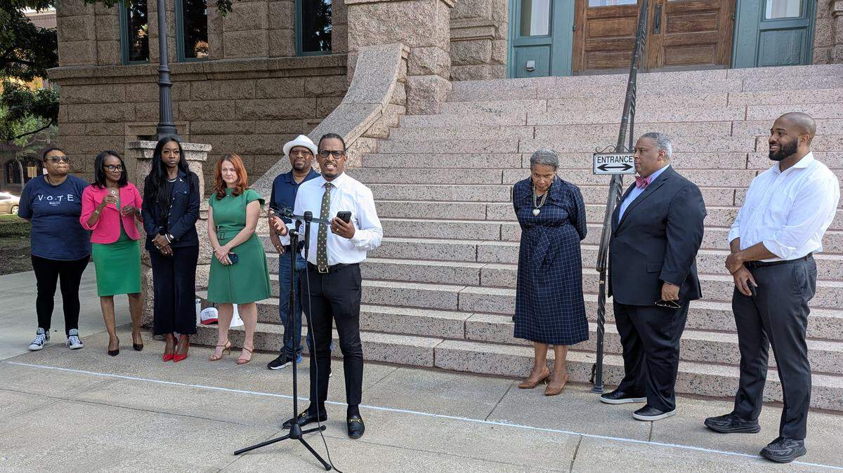 A man standing in front of a microphone surrounded by eight other people in front of a county courthouse building