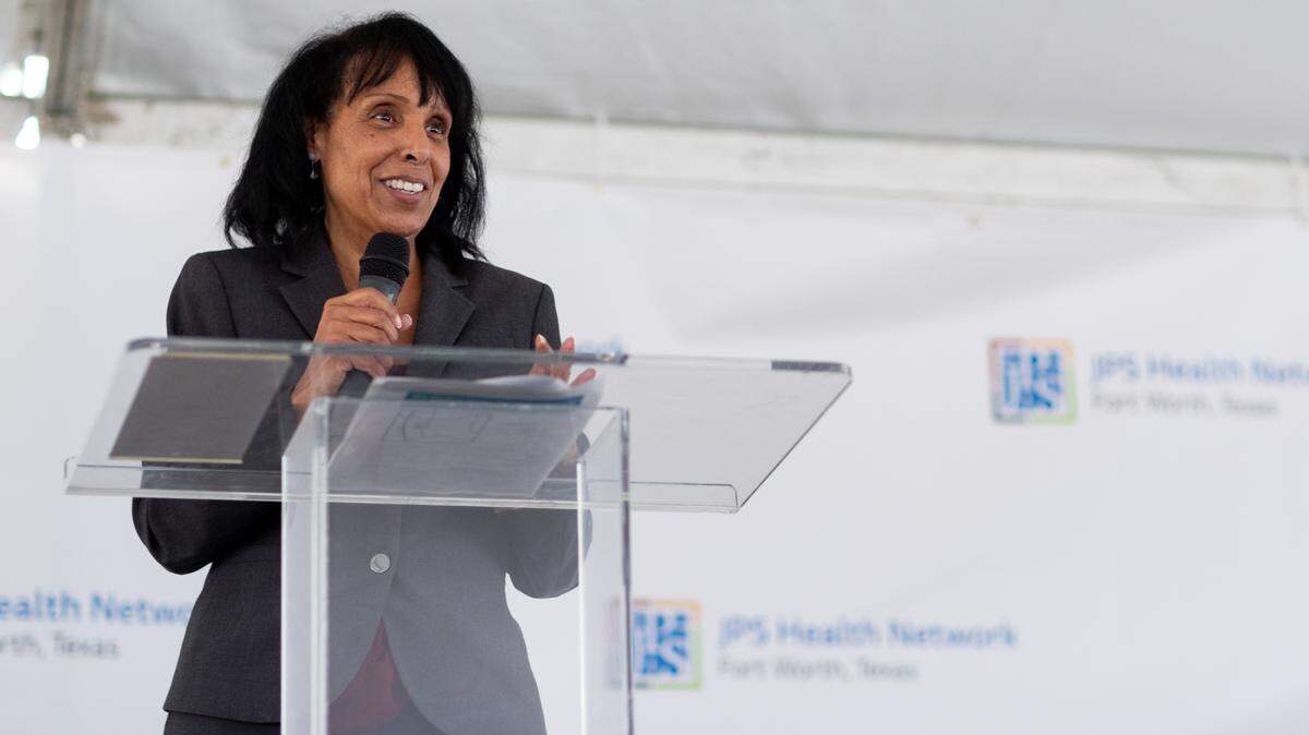 Dr. Duncan stands at a glass podium holding a microphone while smiling while delivering her remarks at the medical facility groundbreaking.