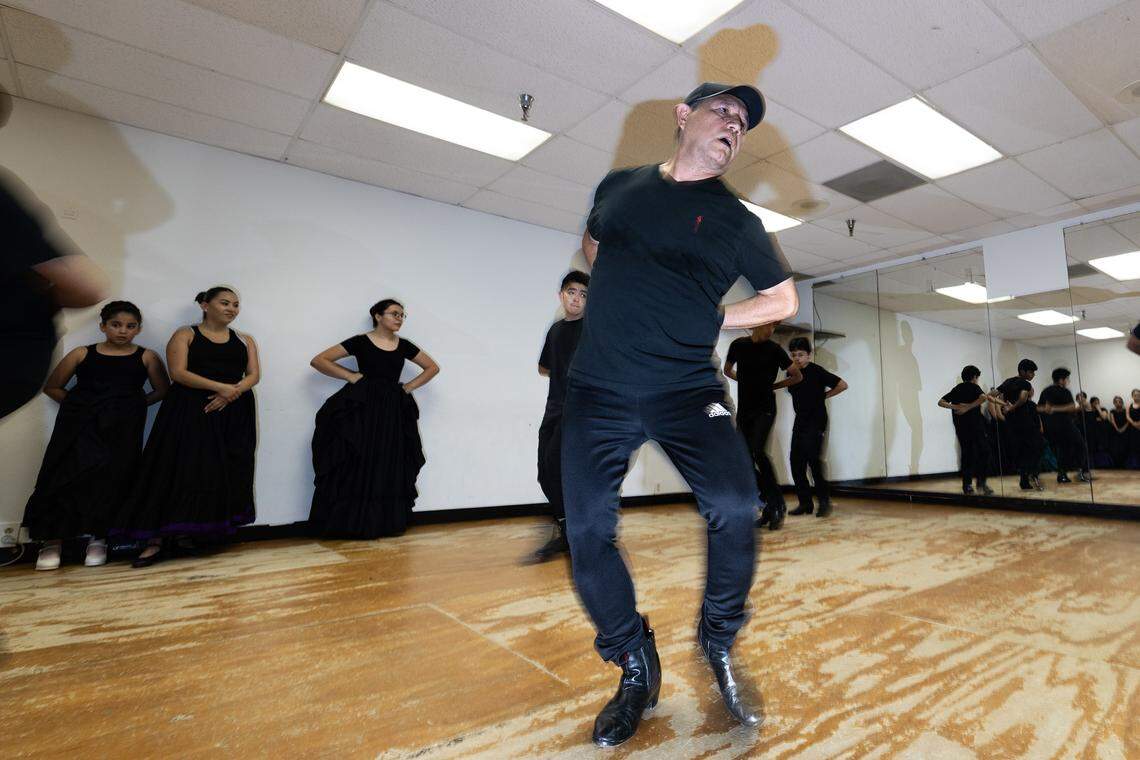 A man all in black dances while in the background more people in black sand at the wall.
