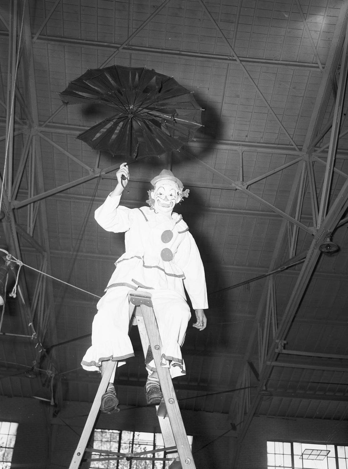 Mr. Hays is shown wearing a clown costume, holding a broken umbrella and standing on a ladder. 