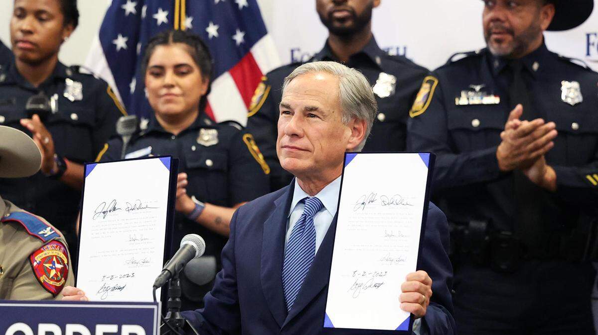 Governor Greg Abbott, sitting at a table, holds a bill in each hand while police officers applaud behind him. 