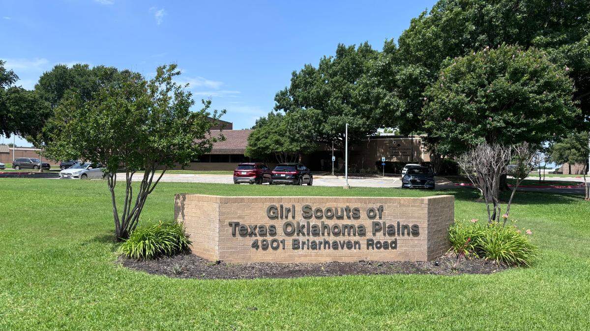 A low brick wall with brown lettering stands in a green lawn. Two small shrub trees flank the sign and a brick office building stands in the background beyond a small parking lot.