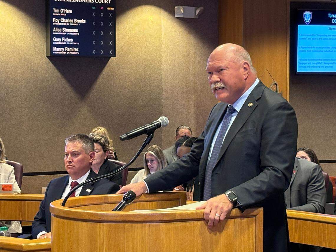 A large man with a mostly bald head and a bushy white mustache stands behind a lectern. His hands rest on the podium. He is wearing a dark grey suit with a navy blue tie. 