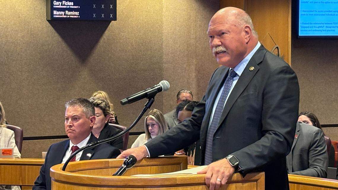 A large man with a mostly bald head and a bushy white mustache stands behind a lectern. His hands rest on the podium. He is wearing a dark grey suit with a navy blue tie. 
