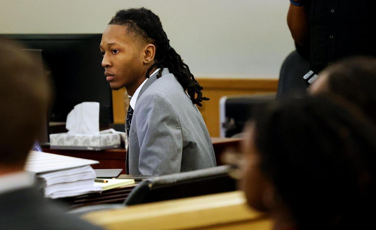 A young man in a grey suite leans forward while listening at the defense table in a crowded courtroom. 