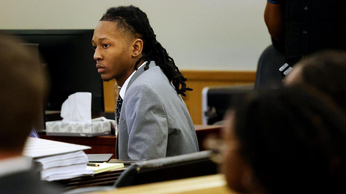 A young man in a grey suite leans forward while listening at the defense table in a crowded courtroom. 