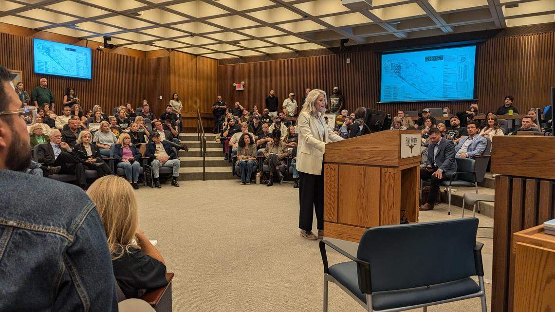 A platinum blond lady speaking at a podium in a government chamber surrounded by onlookers