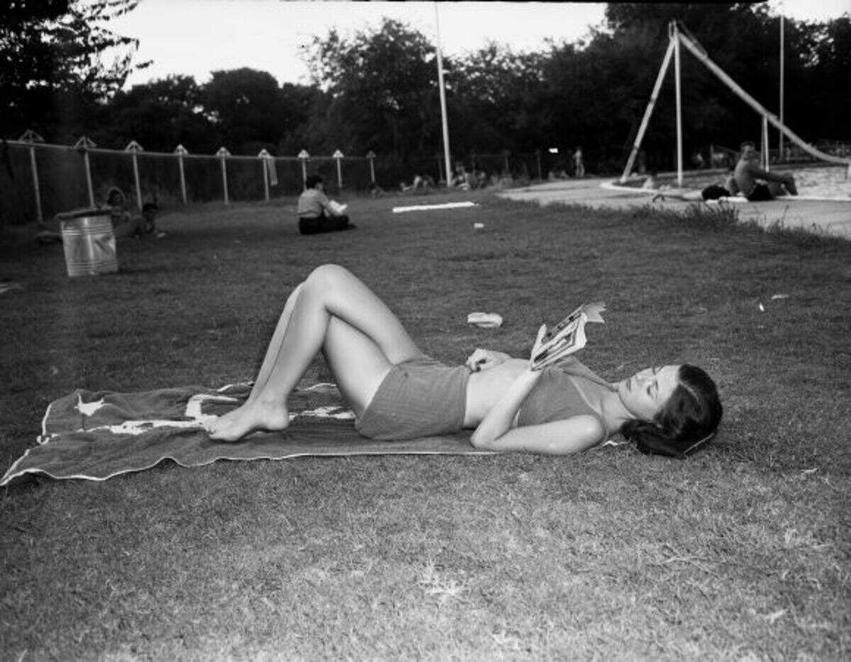 Miss Kenyan is shown laying on a towel on the grass with her legs crossed. She is wearing a pair of shorts and a bikini top as she relaxes and reads a book. 