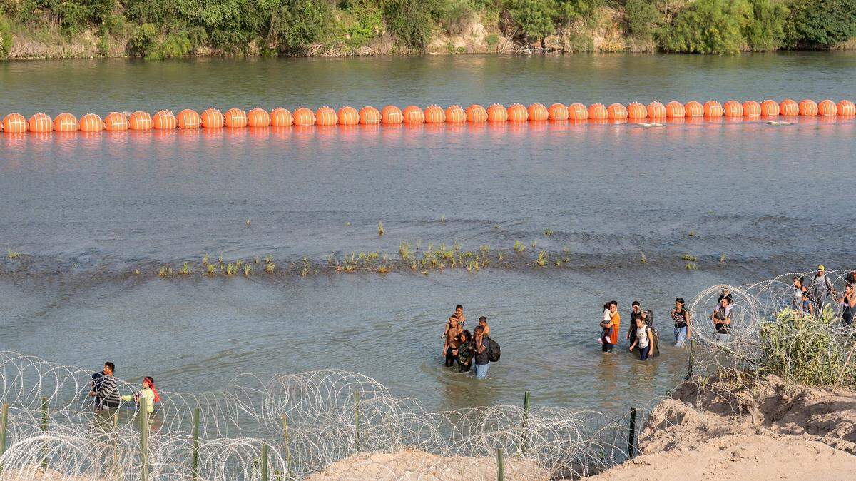 Migrants walk by a string of buoys placed on the water along the Rio Grande border with Mexico in Eagle Pass, Texas, on July 16, 2023. The buoy installation is part of an operation Texas is pursuing to secure its borders, but activists and some legislators say Texas Gov. Greg Abbott is exceeding his authority. (Suzanne Cordeiro/AFP/Getty Images/TNS)